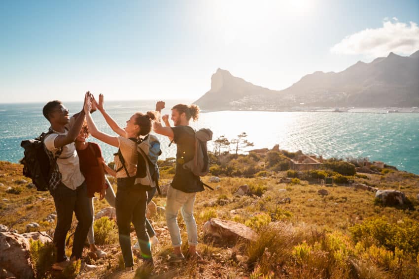 Young adult friends on a hike celebrate reaching a summit near the coast, full length, side view